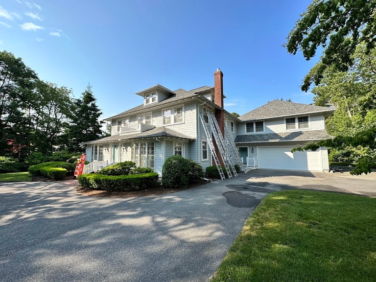 Large two-story white colonial house with attached garage and chimney, surrounded by green lawn and trees on a sunny day