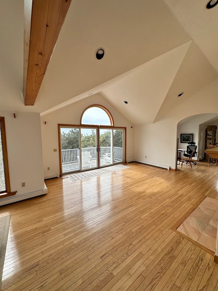 Spacious open loft-style room with wooden floors, vaulted ceiling, exposed beam, and glass doors leading to a deck with forest views