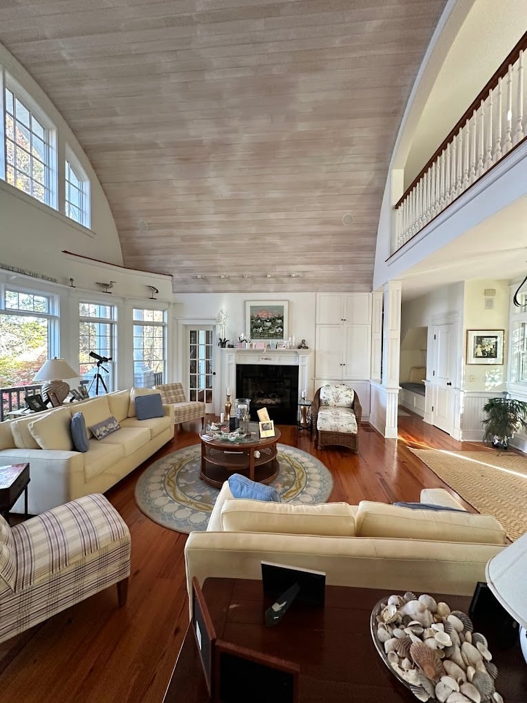 Bright open-plan living room with cream seating, wooden floors, fireplace, vaulted ceiling, and large windows with natural light